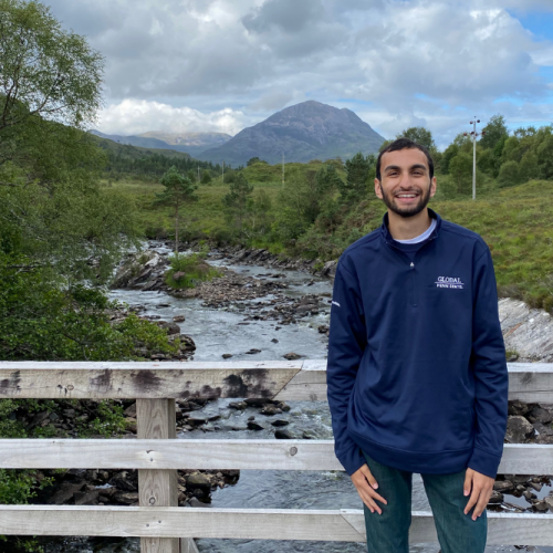 Psychology and Labor and Human Resources student, Nicholas Banerjee, stands on a wooden bridge in front of a rocky stream. Trees and greenery surround the stream, and mountains and billowing clouds are visible in the distance. The picture was taken in Scotland where Nicholas conducted workplace behavioral research as a Paterno Fellow.
