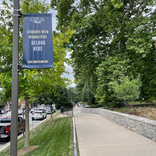 A blue "Students with Disabilities Belong Here" UPUA banner hangs above a sidewalk along East College Avenue on the University Park campus.