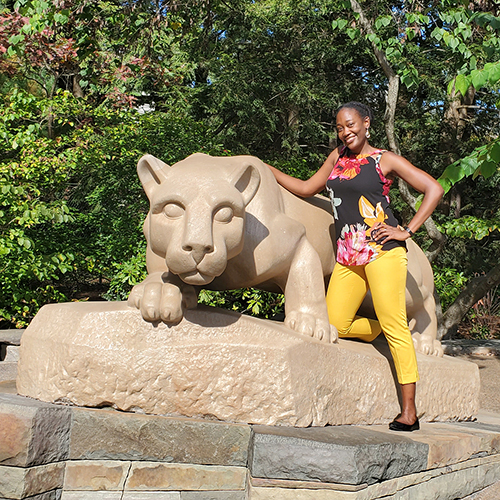 African American woman with yellow pants and flowery top standing by Penn State Nittany Lion Shrine.